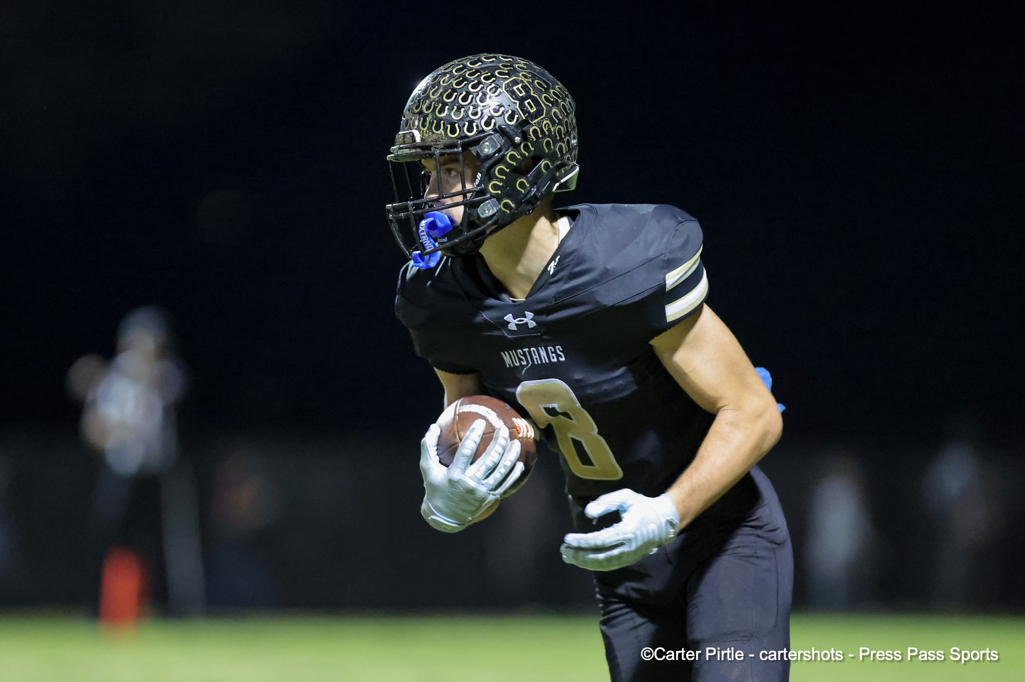 Football goalie in a black 'Mustangs' jersey runs with the ball at night, wearing a patterned helmet and gloves.