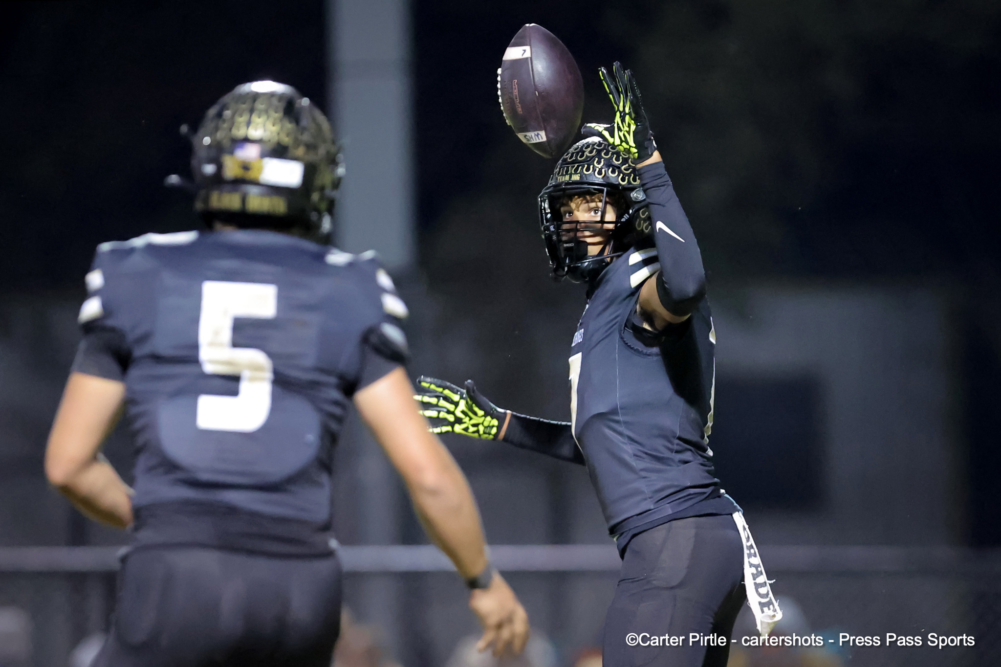 A football player in a navy uniform reaches up to catch a spiraling ball at night, another player seen in the foreground.