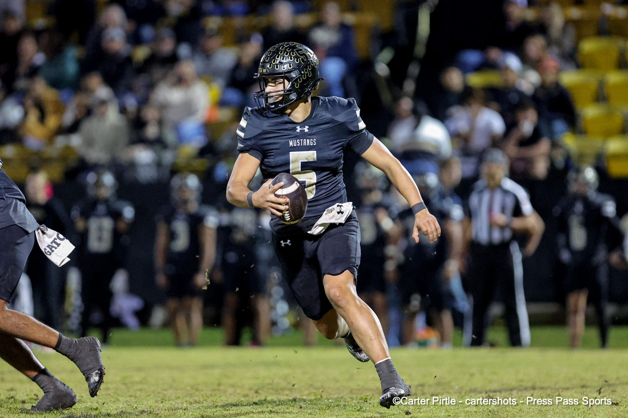 Football player in a dark uniform carrying a ball, running on the field with a crowd in the stands behind him, helmet on.