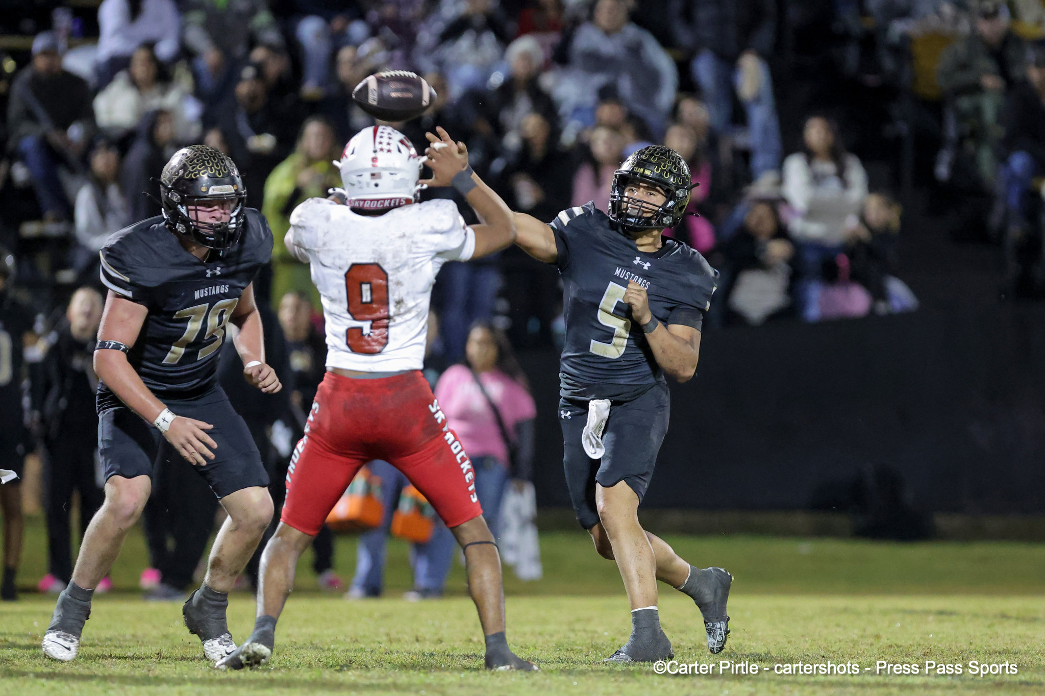 Quarterback in a white jersey throws a football while two defenders in dark Mustangs uniforms rush toward him on a nighttime football field with a cheering crowd in the stands.