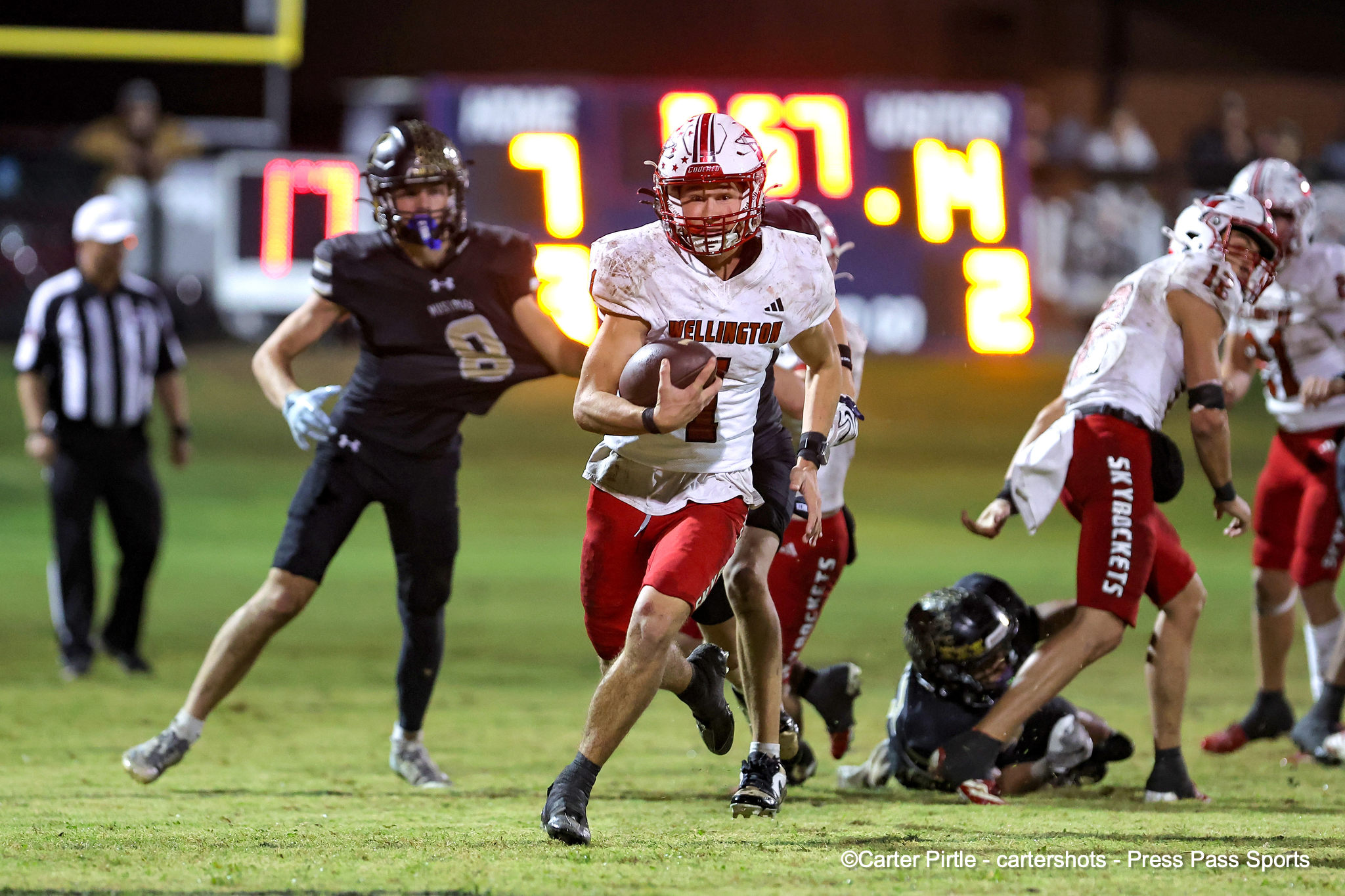 Football player in a white Wellington jersey carries the ball while sprinting down the field as defenders close in during a night game on a grassy field.