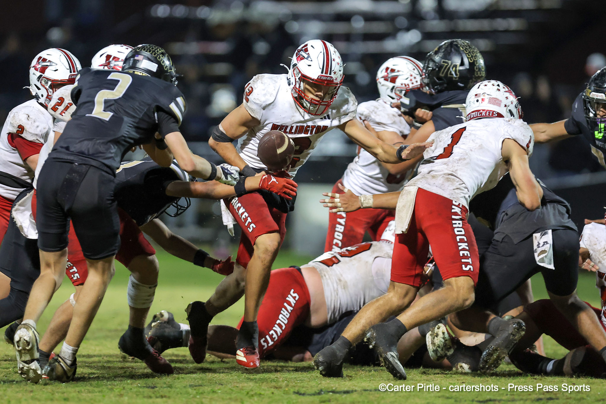 Wellington football player in white jersey carrying the ball, sprinting through a cluster of defenders in red, white, and black uniforms on a muddy field.
