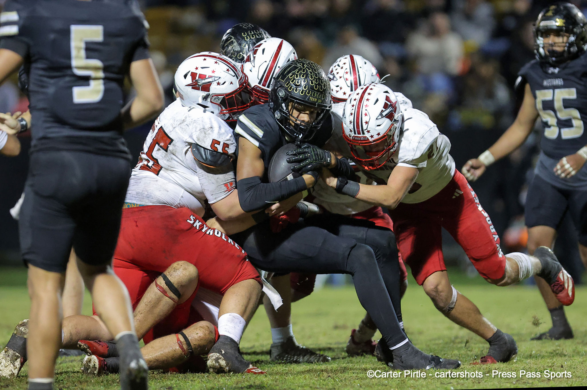 Players in dark uniforms converge on a ball carrier as teammates in white jerseys tackle him near the line of scrimmage.