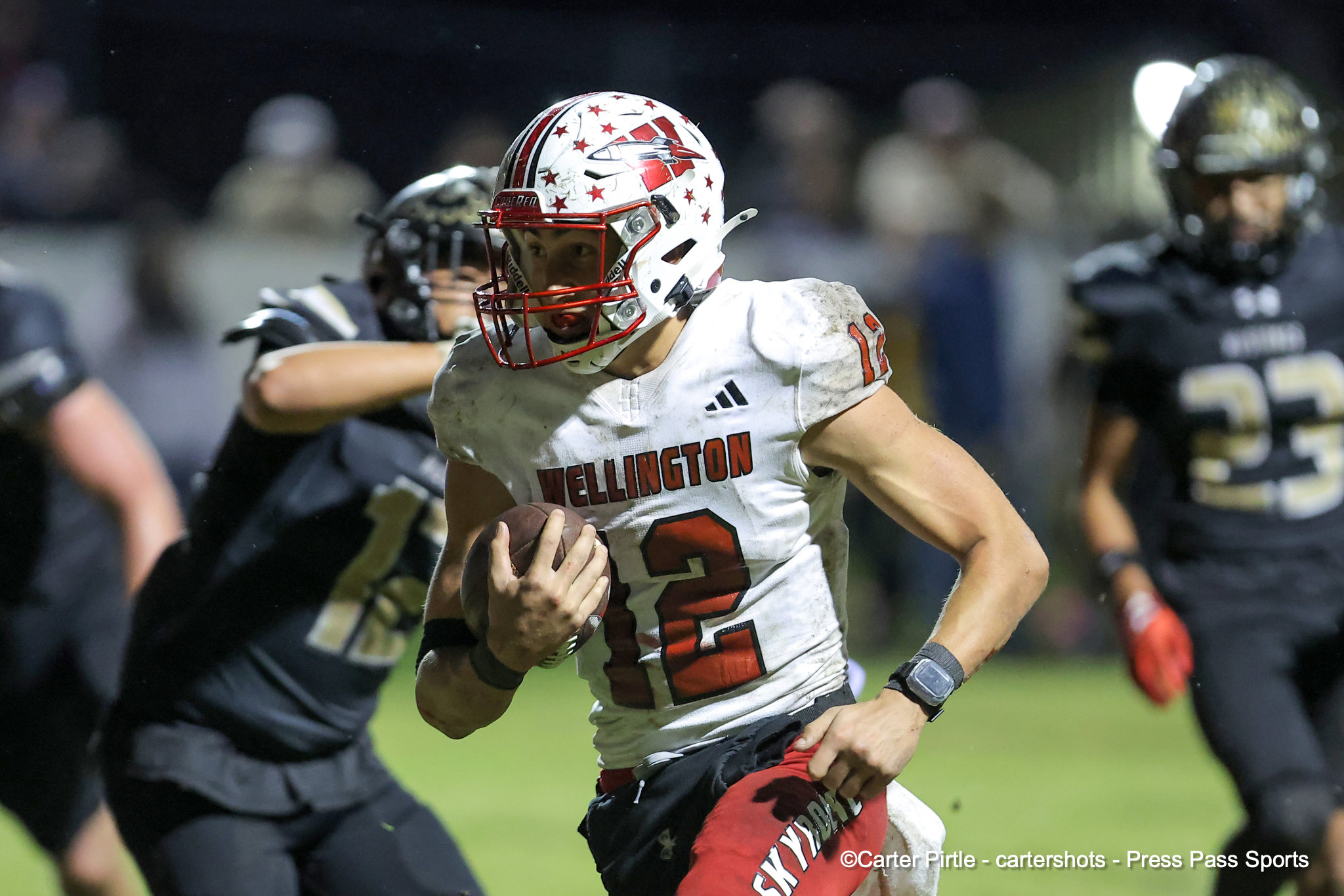 Football player in white with red helmet and stars, carrying the ball during a night game amid defenders.