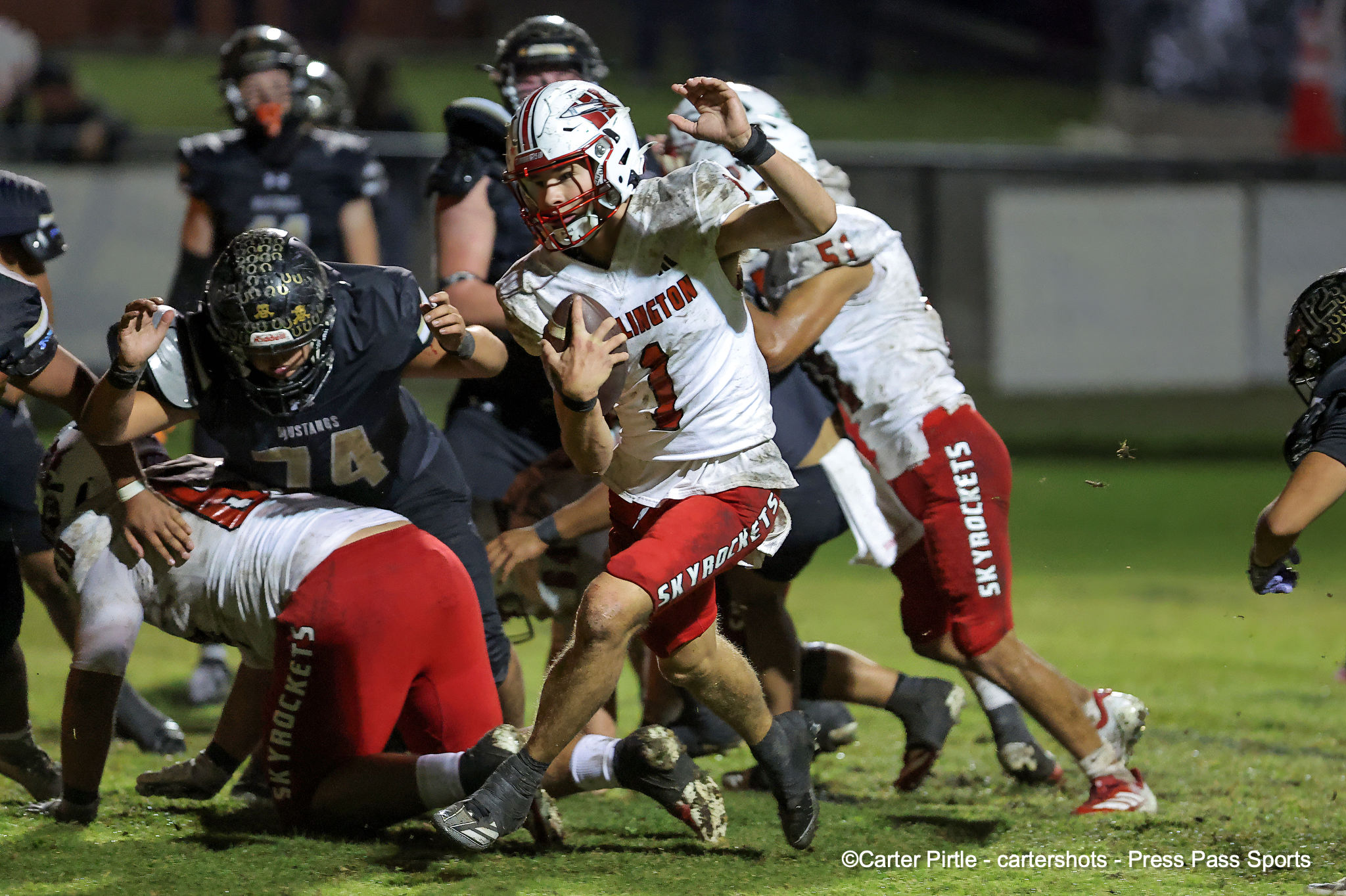 Football players in white and red uniforms collide on a muddy field as a ball carrier runs forward and others pursue.
