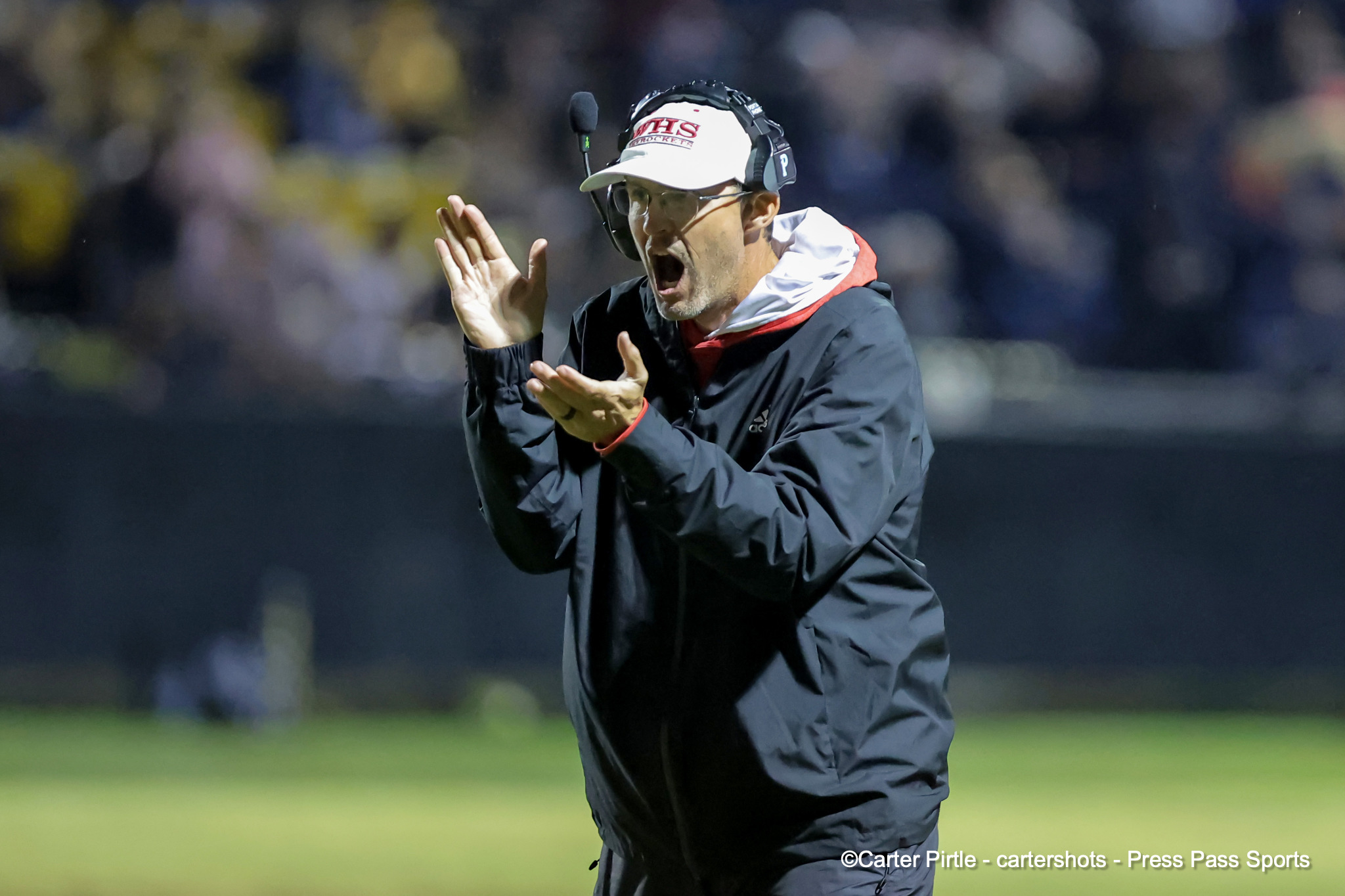 Coach or staff member in a headset shouting with hands raised during a night game on a field.
