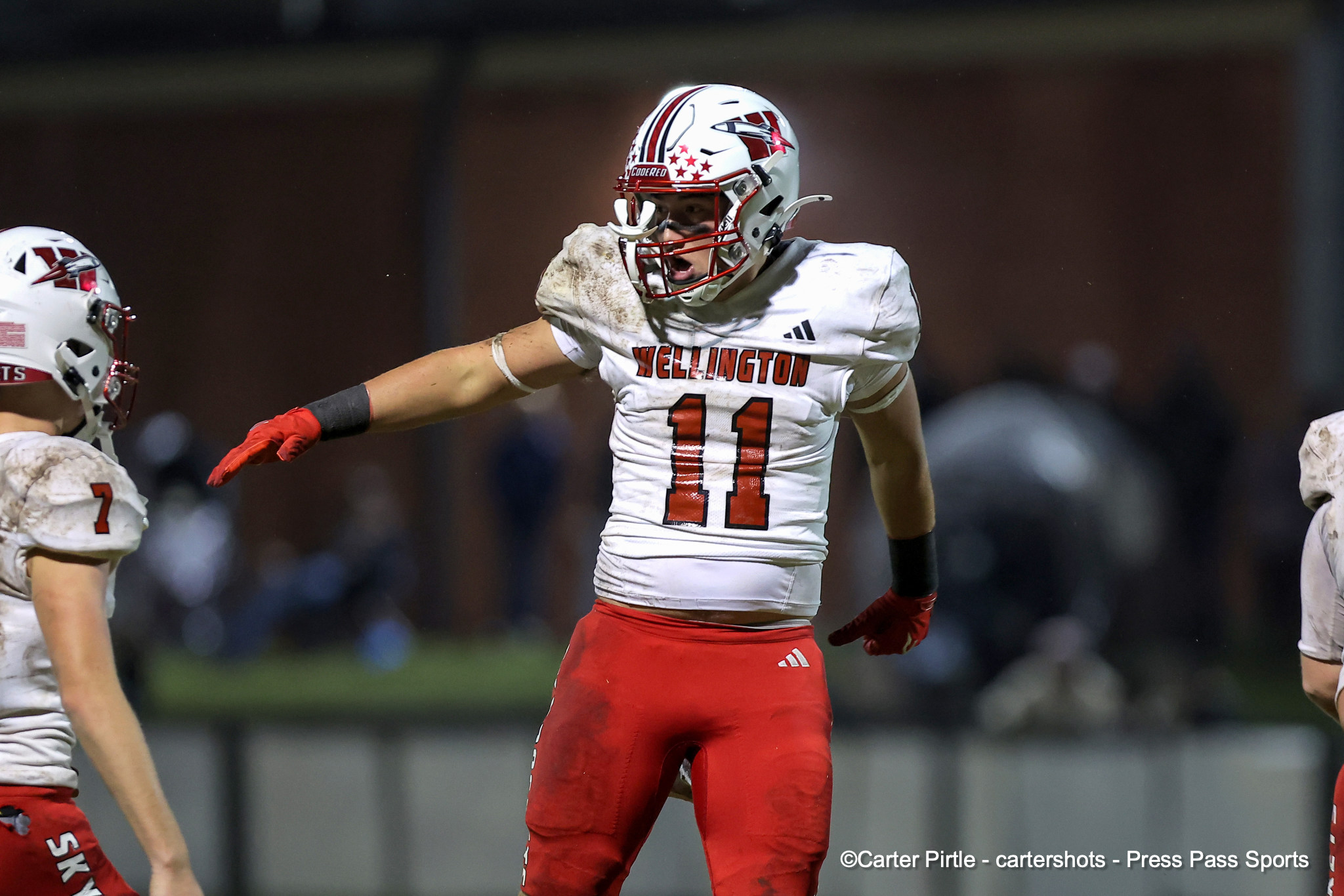 Football player wearing Wellington #11 in white jersey and red pants, celebrating with arms outstretched on the field.