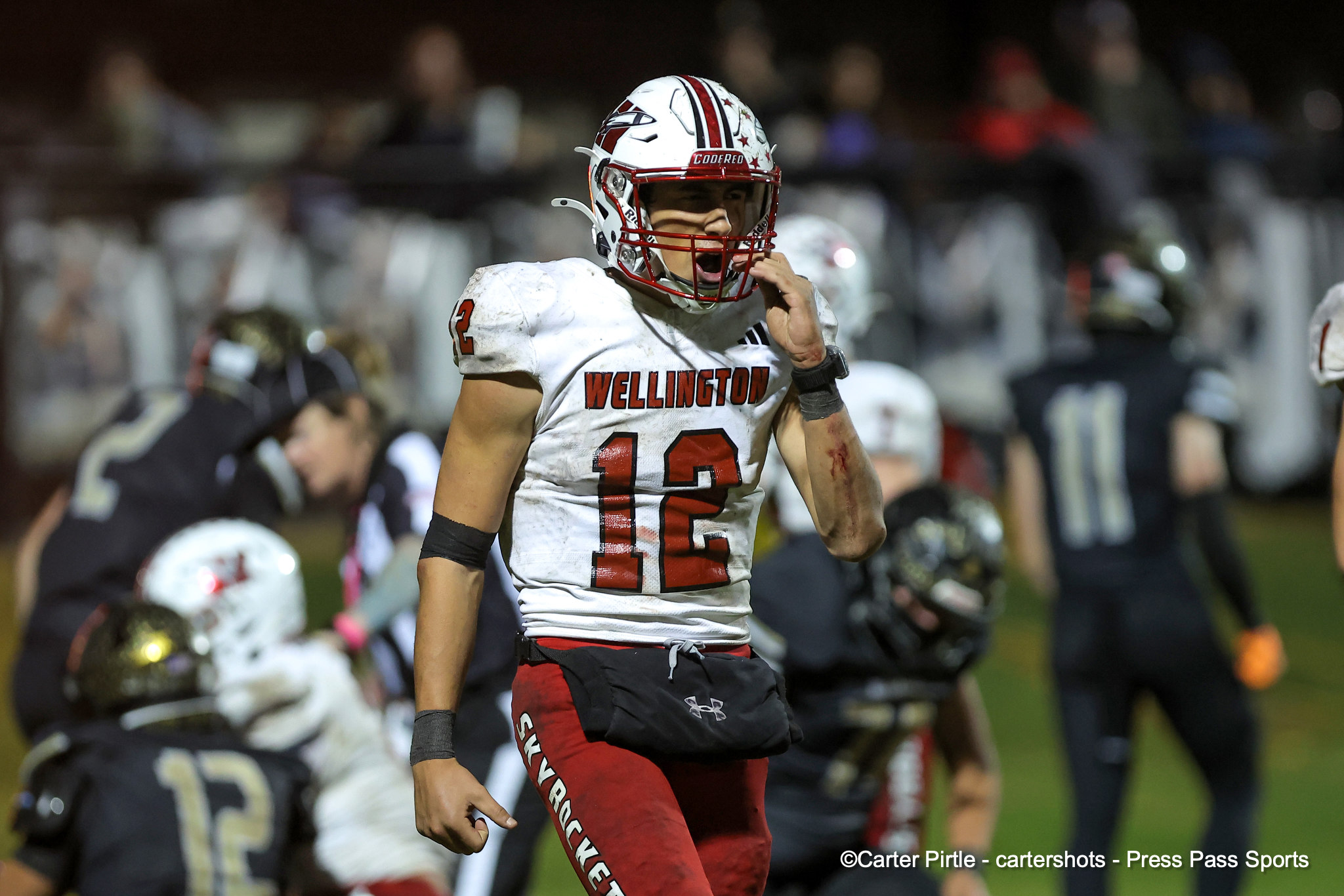 Football player in white Wellington jersey with number 12, helmet, on a night game with blurred crowd in background.