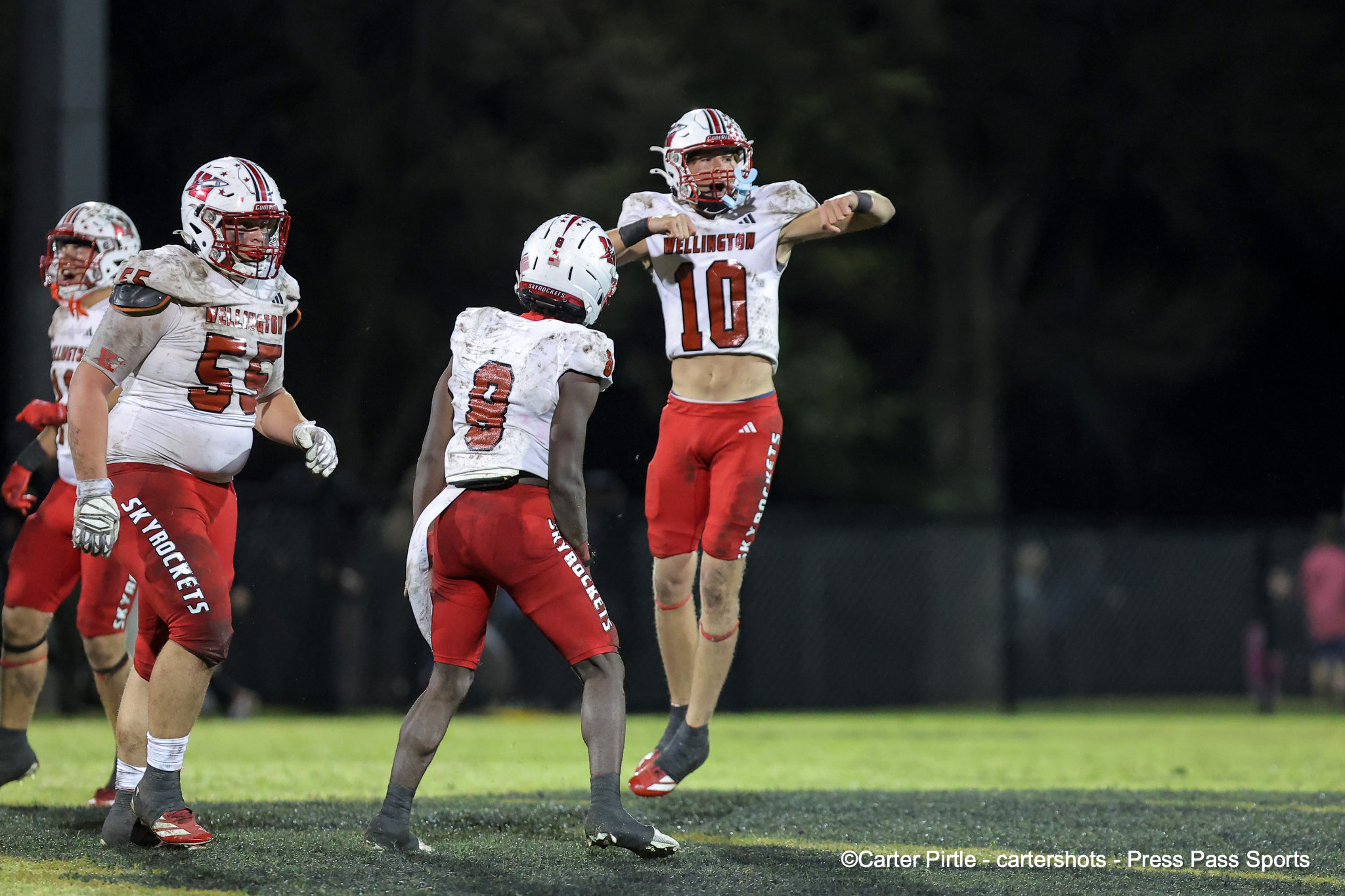 Group of football players in white and red uniforms celebrate on a muddy, well-lit field at night, with one jumper raising his arms in triumph.