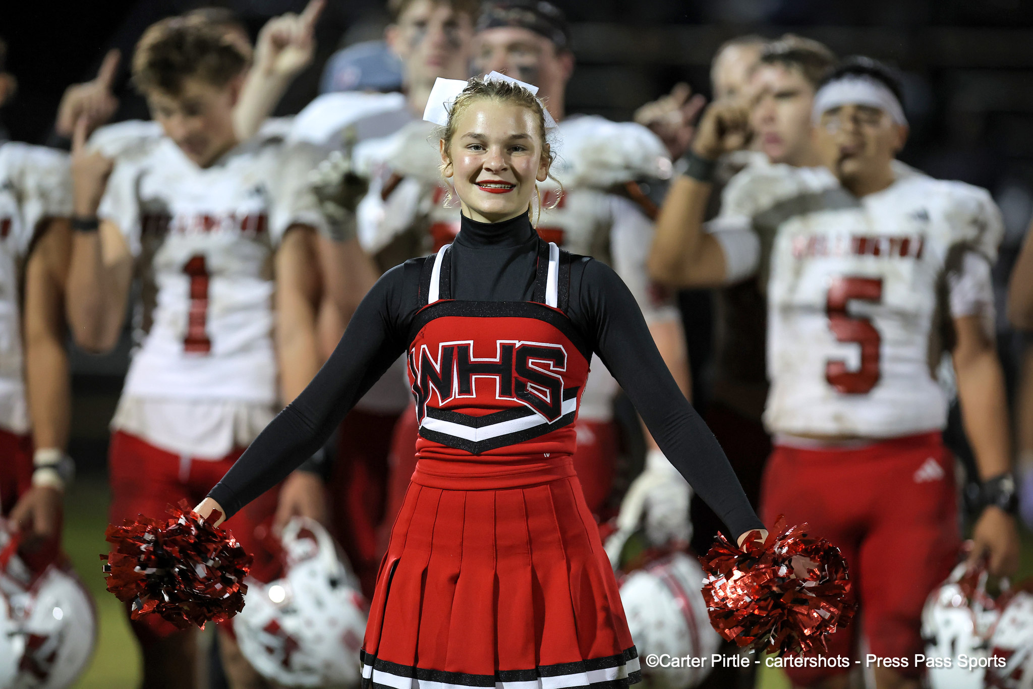 Cheerleader in a red and black uniform smiles at the camera, holding red pom-poms with teammates blurred in the background at a game.