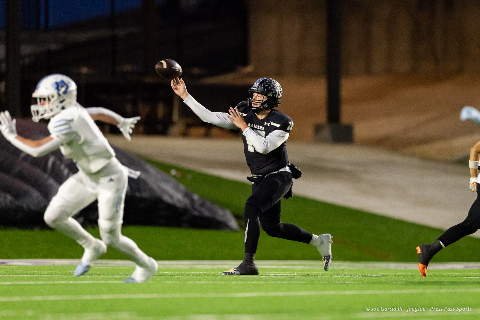 west-plains-vs-randall-2026-jpegJoe-pps-2 | Press Pass Sports Raiders quarterback in a black uniform throws a pass during a night game, with a pursuing white-uniform defender nearby.
