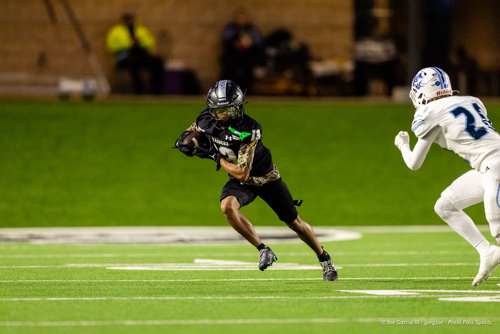 west-plains-vs-randall-2026-jpegJoe-pps-3 | Press Pass Sports Football player in black with camo sleeves running with the ball as a defender in white closes in on a night-lit green turf field.