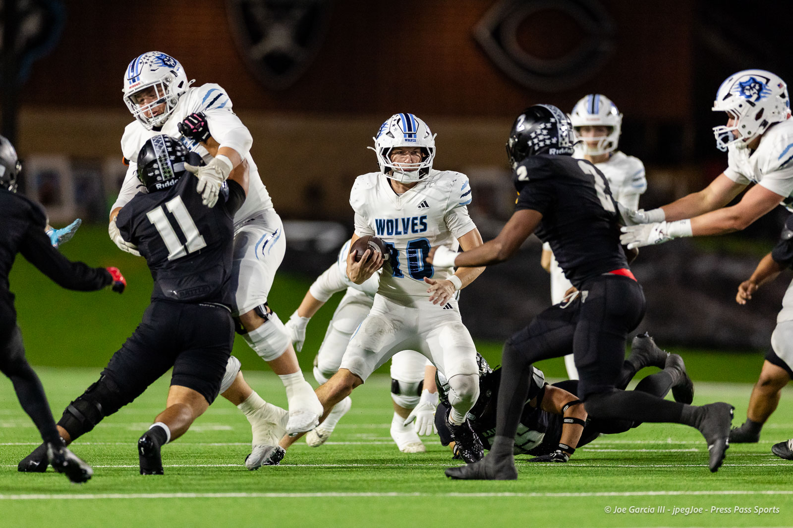 west-plains-vs-randall-2026-jpegJoe-pps-9 | Press Pass Sports Wolves quarterback #10 in white uniform holding the ball and looking for a path as defenders in black close in during a night game on a bright green field.