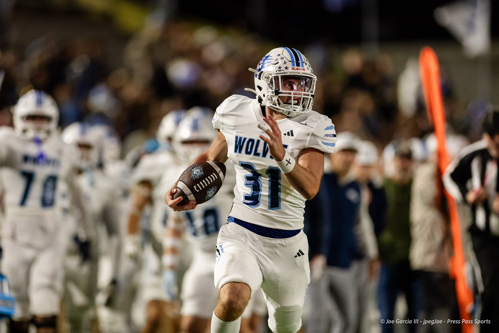 west-plains-vs-randall-2026-jpegJoe-pps-14 | Press Pass Sports American football player in a white and blue uniform (number 31) runs with the ball during a game, crowd in the background