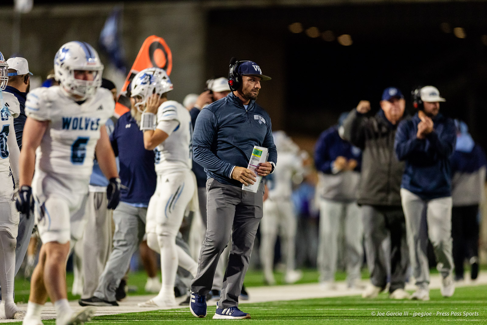 west-plains-vs-randall-2026-jpegJoe-pps-16 | Press Pass Sports Coach in navy pullover and headset walking on the sideline, holding a clipboard.