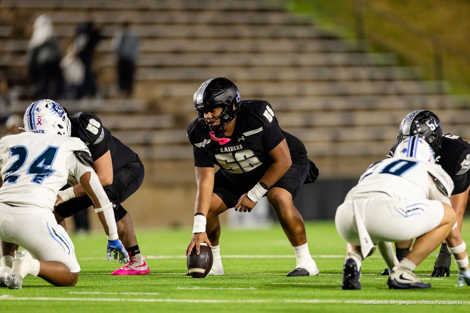 west-plains-vs-randall-2026-jpegJoe-pps-19 | Press Pass Sports Raiders offensive lineman in black #56 crouches over the ball at the line of scrimmage, ready to snap as white-clad defenders approach on a bright field.