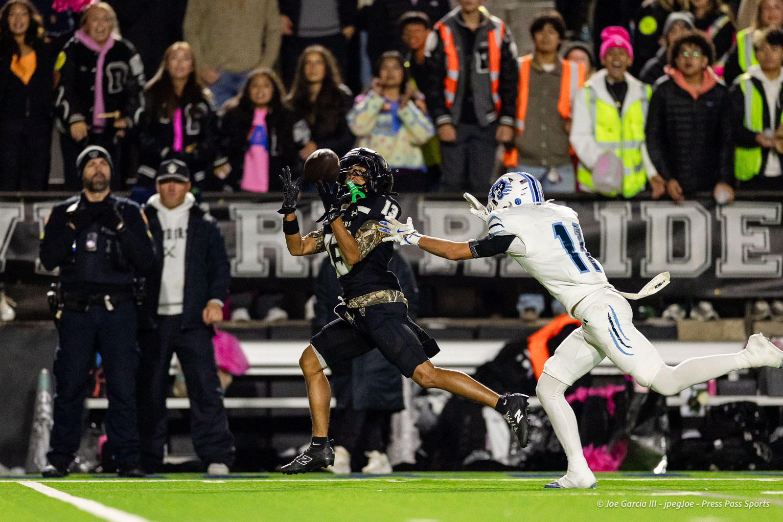Football player in black jumps to catch the ball as a white-uniform defender reaches in front of a cheering crowd behind.]
