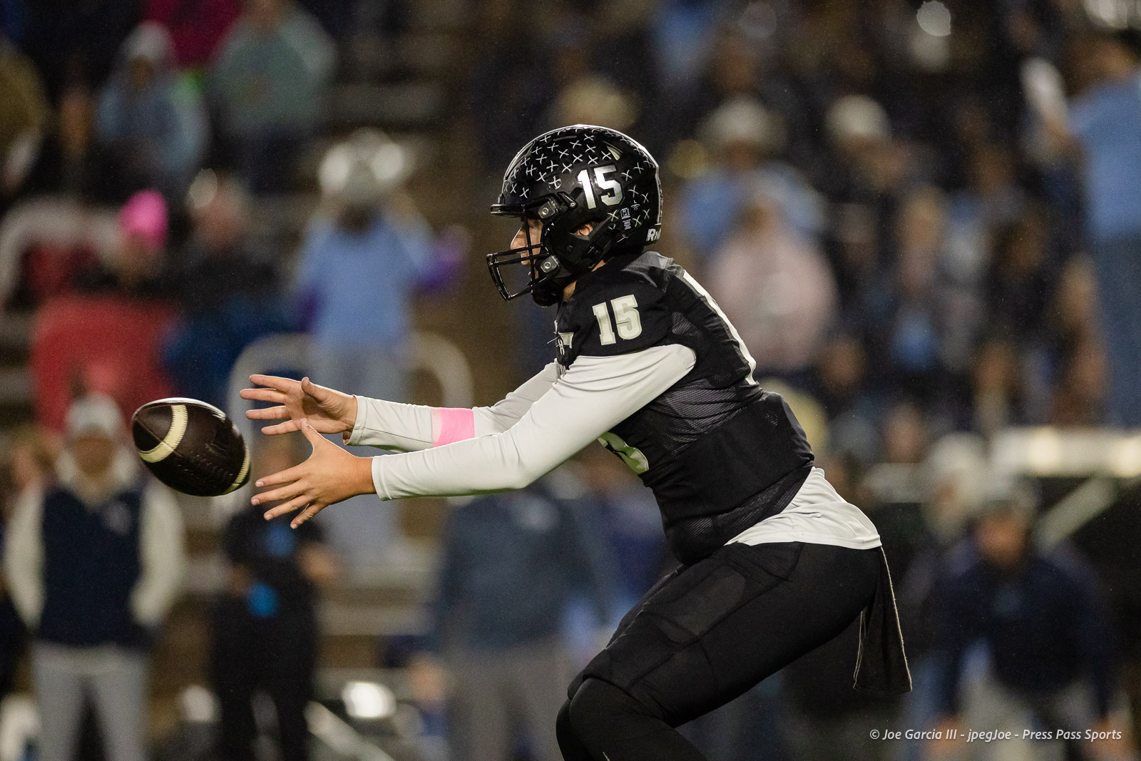 west-plains-vs-randall-2026-jpegJoe-pps-24 | Press Pass Sports American football player in a black uniform and helmet (number 15) reaches to catch a ball with a blurred crowd in the background, mid-action symmetry.