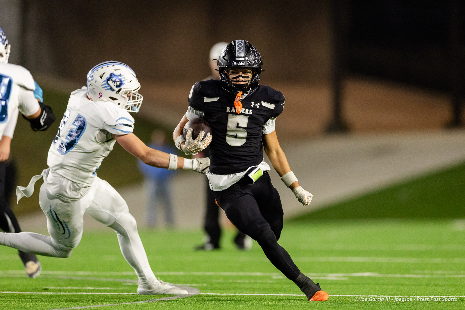 west-plains-vs-randall-2026-jpegJoe-pps-25 | Press Pass Sports Football player in black uniform (#5) Raiders carries the ball, pursued by a white-jersey defender on a night game on a turf field.