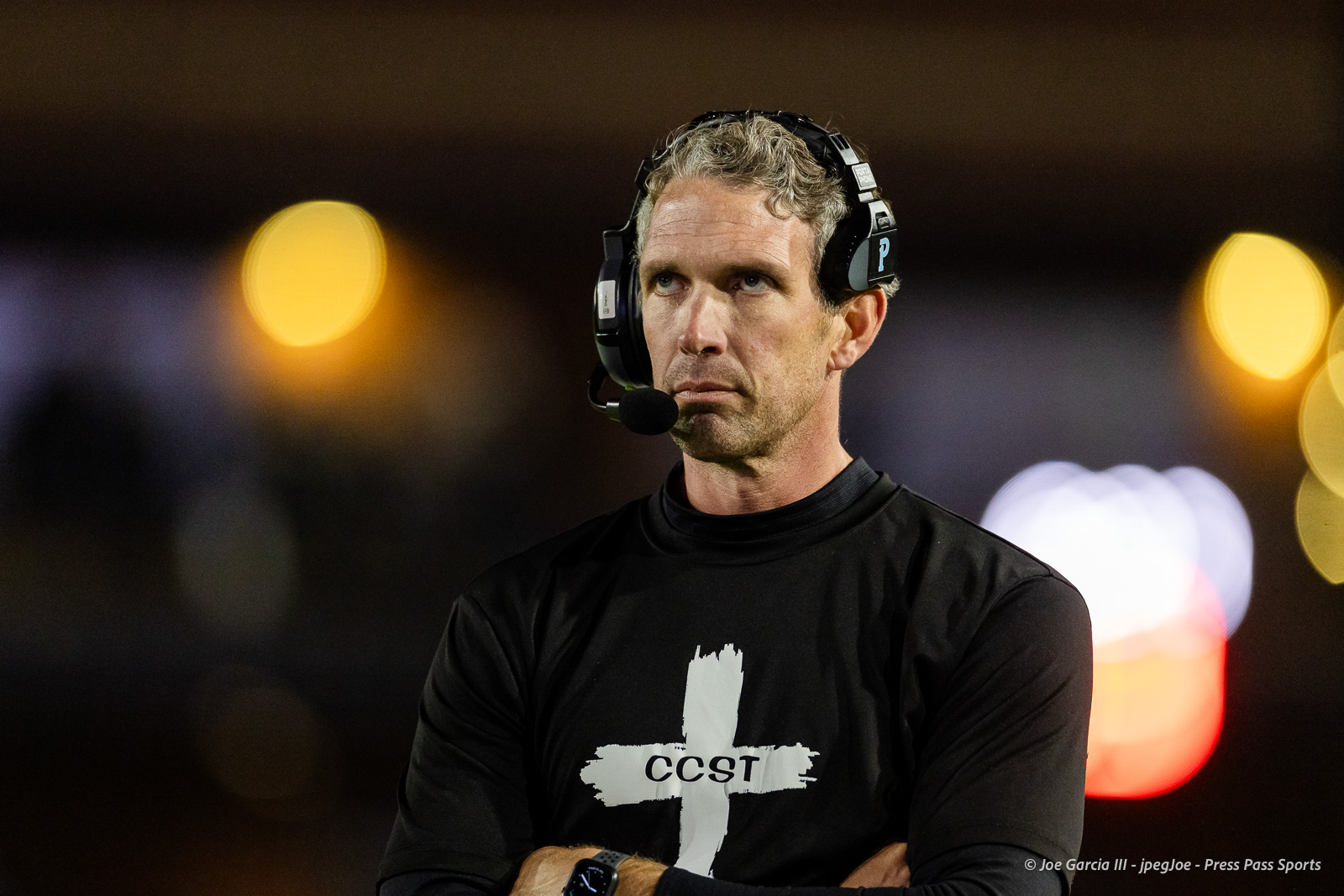 west-plains-vs-randall-2026-jpegJoe-pps-26 | Press Pass Sports Man wearing a headset with microphone and a black CCST shirt, arms crossed, at night with blurred city lights in the background.