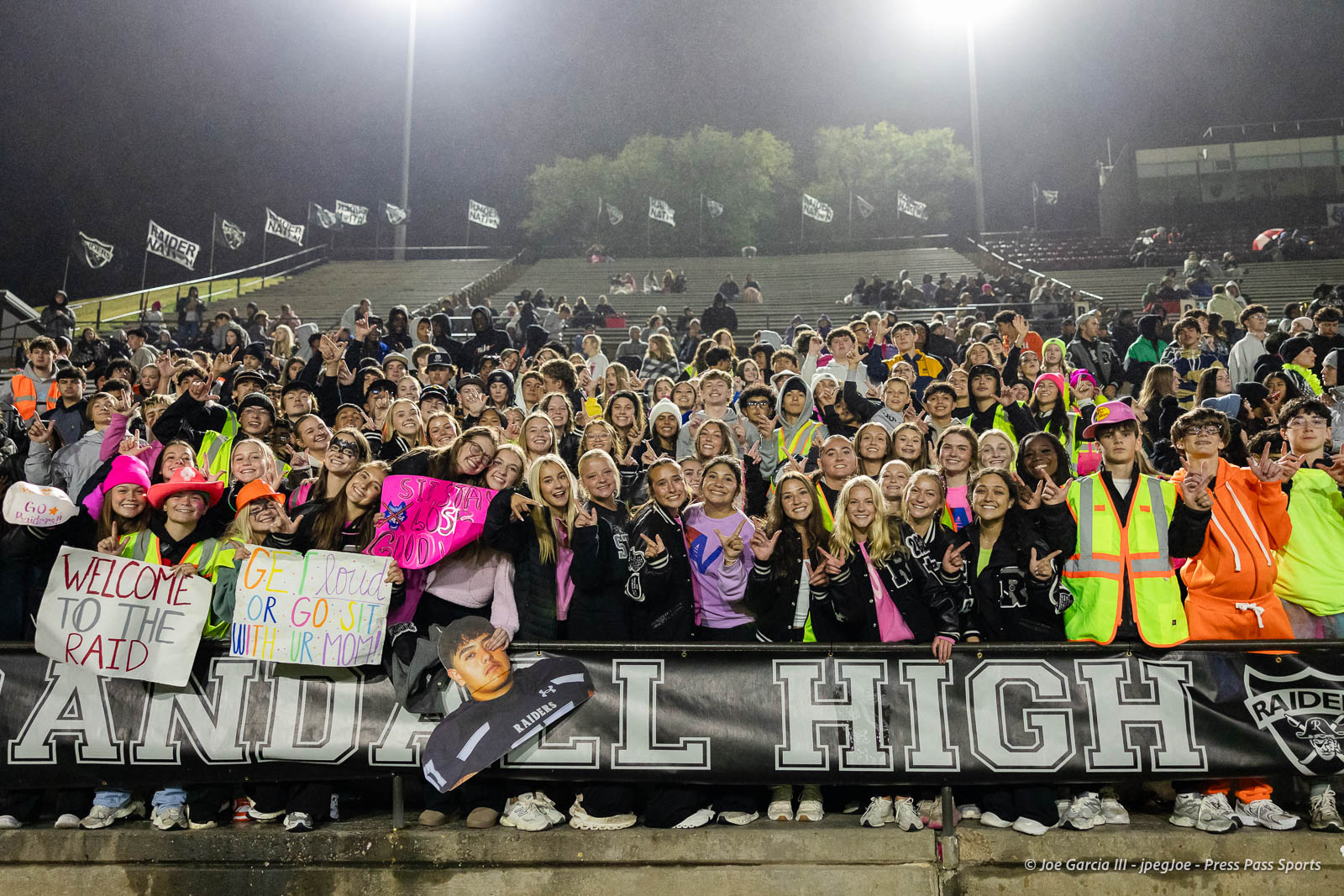 west-plains-vs-randall-2026-jpegJoe-pps-28 | Press Pass Sports Large group of students at a nighttime stadium rally, holding colorful signs and wearing bright vests, behind a Raiders banner.