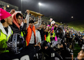 west-plains-vs-randall-2026-jpegJoe-pps-29 | Press Pass Sports Nighttime football crowd on the sideline cheering as a player lifts a brown plaque or banner high above the barrier in triumph.