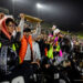 west-plains-vs-randall-2026-jpegJoe-pps-29 | Press Pass Sports Nighttime football crowd on the sideline cheering as a player lifts a brown plaque or banner high above the barrier in triumph.