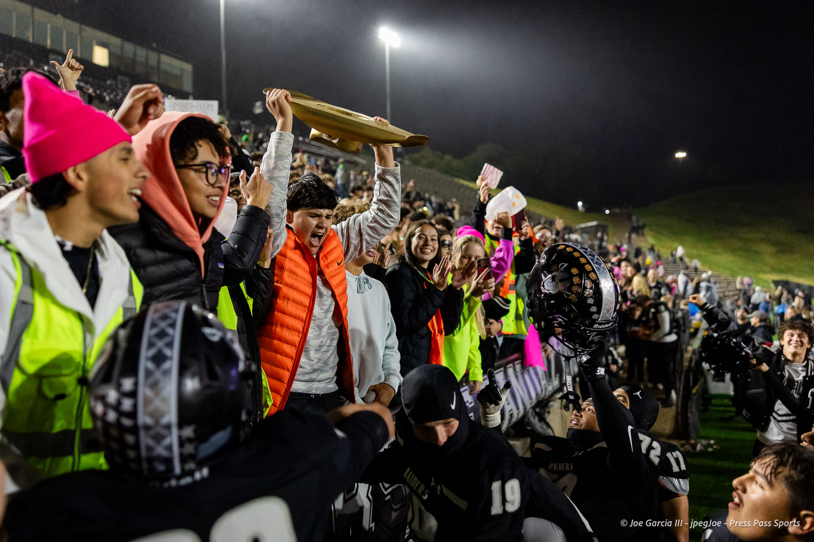 west-plains-vs-randall-2026-jpegJoe-pps-29 | Press Pass Sports Nighttime football crowd on the sideline cheering as a player lifts a brown plaque or banner high above the barrier in triumph.
