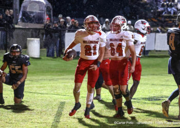 20261030-Wellington-Wheeler-FB-011 | Press Pass Sports Wellington football players in white and red celebrate on a rain-soaked field, one carrying the ball amidst teammates.
