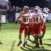 20261030-Wellington-Wheeler-FB-011 | Press Pass Sports Wellington football players in white and red celebrate on a rain-soaked field, one carrying the ball amidst teammates.