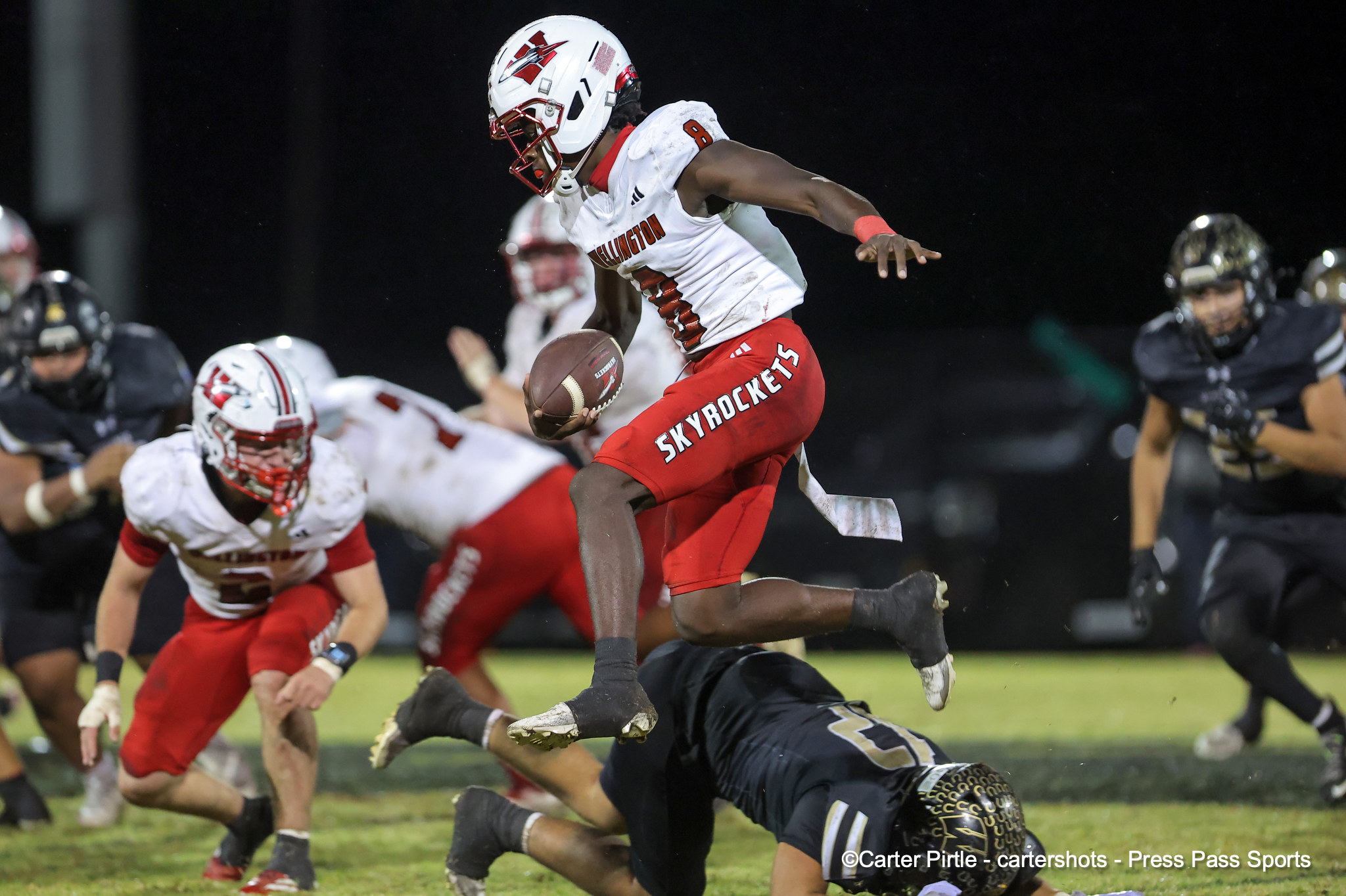 Tall football player in a white and red Illinois Skyrockets uniform leaps with the ball against a night game backdrop, as defenders dive and pursue him.