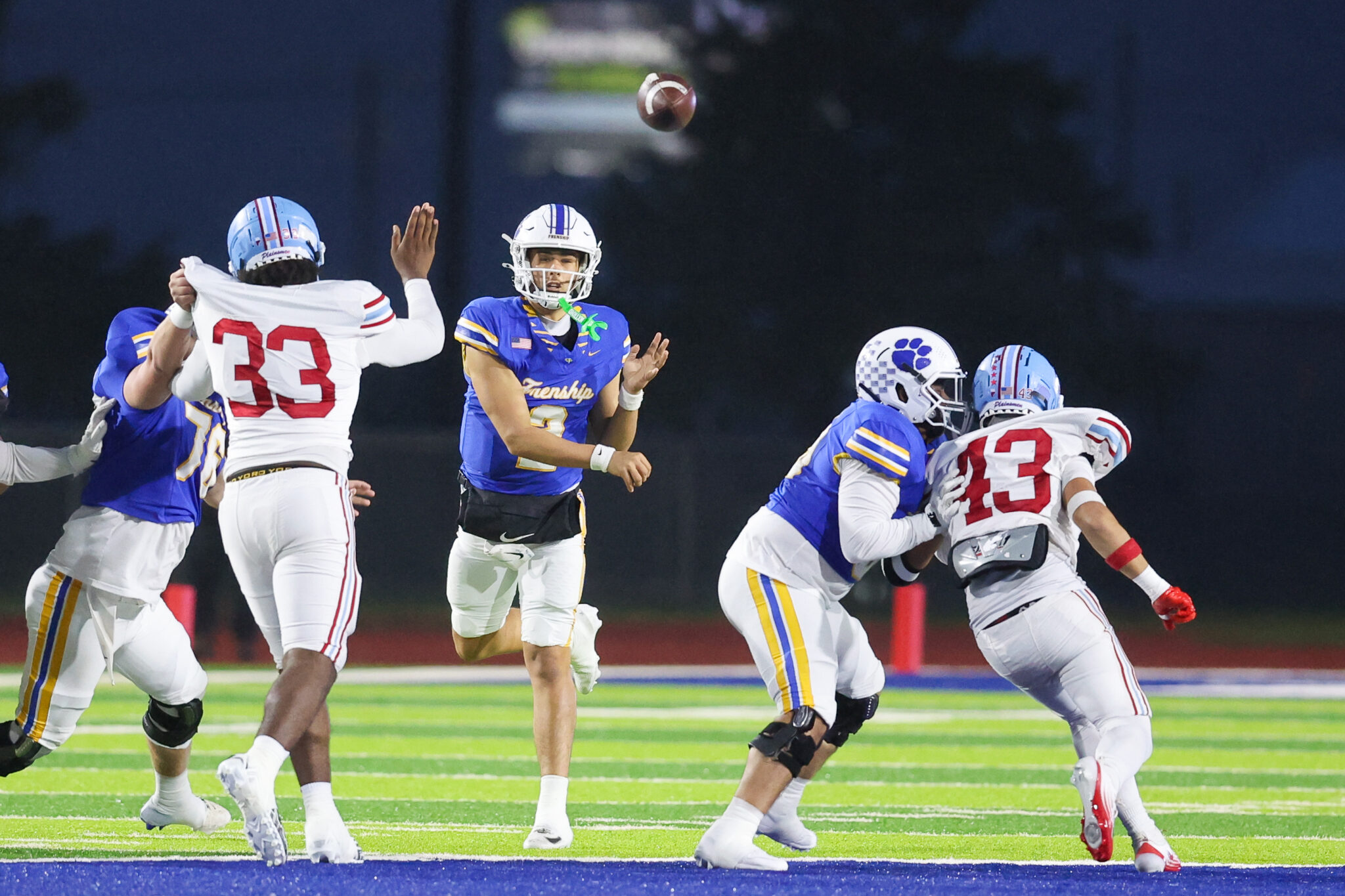 Seabolt Chase-18740 | Press Pass Sports Football players in blue and white uniforms reach for a high ball on a green field at night; quarterback looks up at the pass.