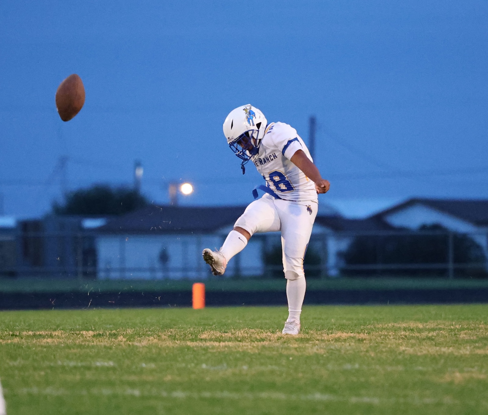 Kicker in white uniform punts a football on a grassy field at dusk, blue sky in the background.