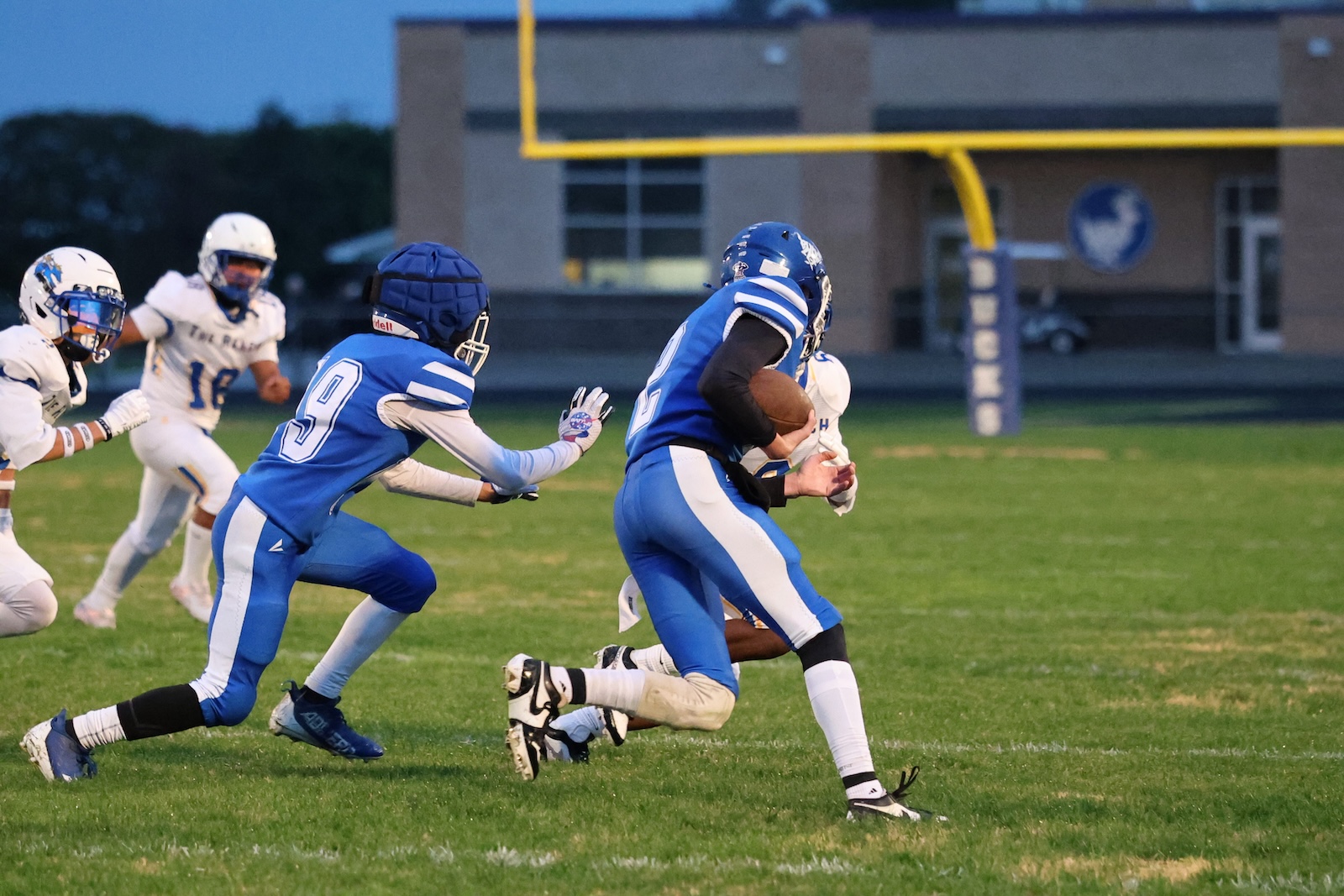 Blue team football player carries the ball and runs forward while a defender in blue pursues from behind on a grassy field, with white-clad players closing in and a goalpost in the background.