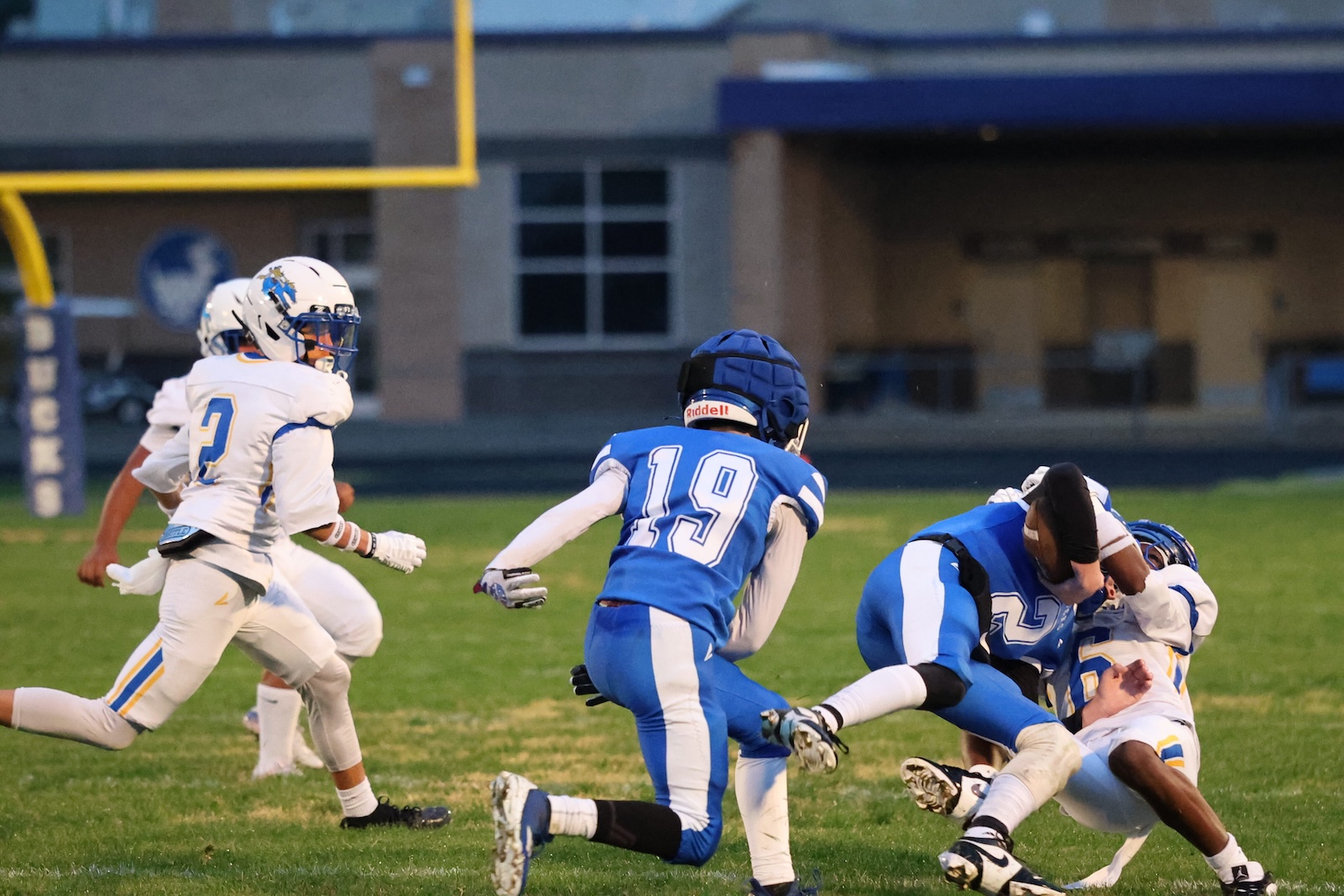 Football players in blue and white uniforms collide on a grassy field during a game, one tackled to the ground.