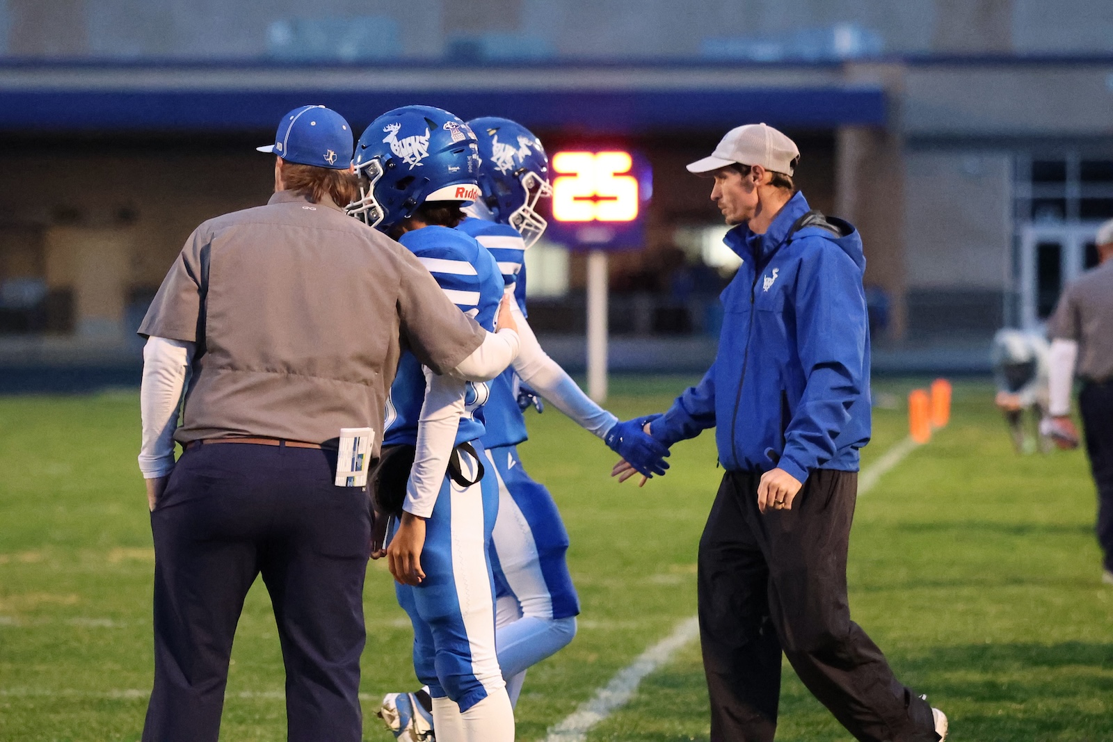 Football players in blue uniforms and helmets shake hands with coaches on the sideline before a game on a grassy field, a glowing scoreboard in the background.