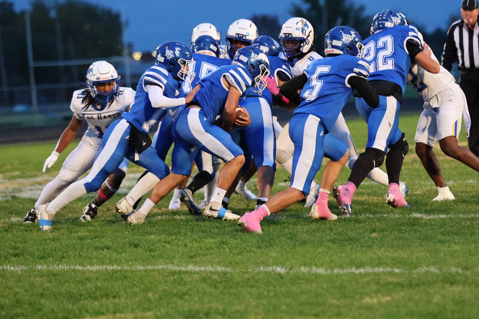 Group of football players in blue jerseys converge around the ball on a grassy field during a play at dusk/faint evening light, jostling for possession.