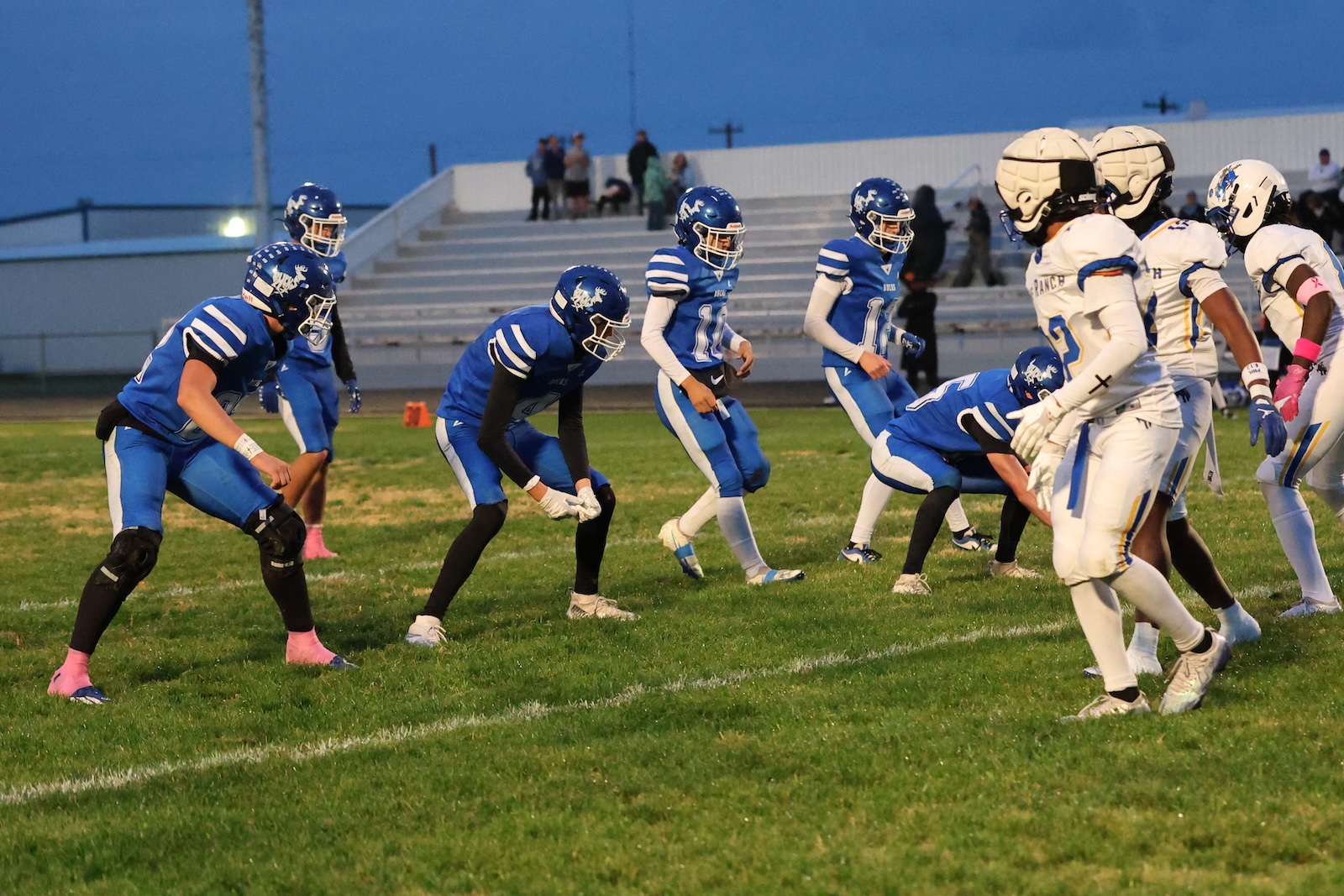 High school football players in blue uniforms line up against white-uniformed opponents on a grassy field at dusk.