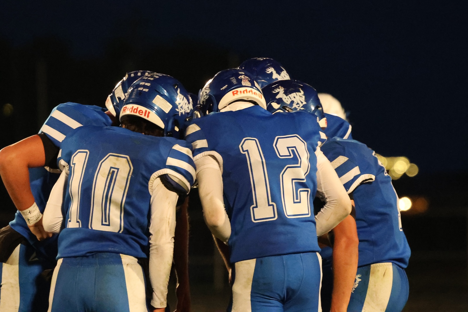 Blue-clad football players huddle with helmets on, numbers 10 and 12 visible on their backs, at night.