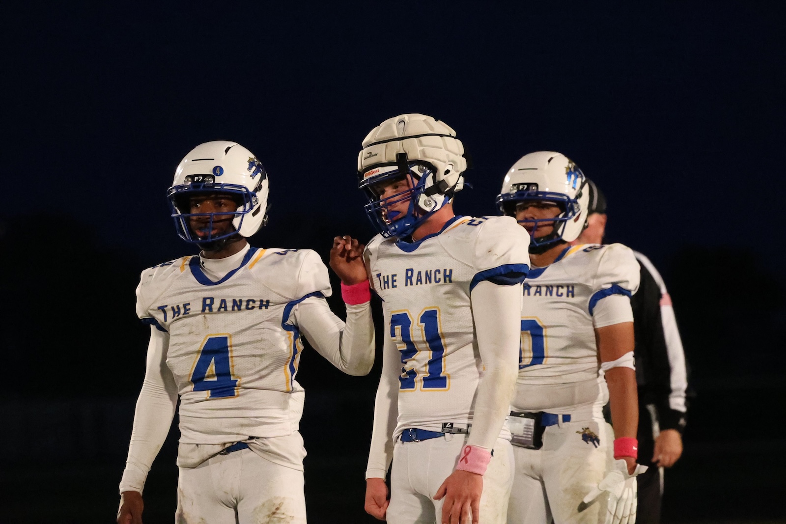 Three football players from The Ranch stand together on the field at night in white uniforms and helmets, with jersey numbers 4 and 31 visible.