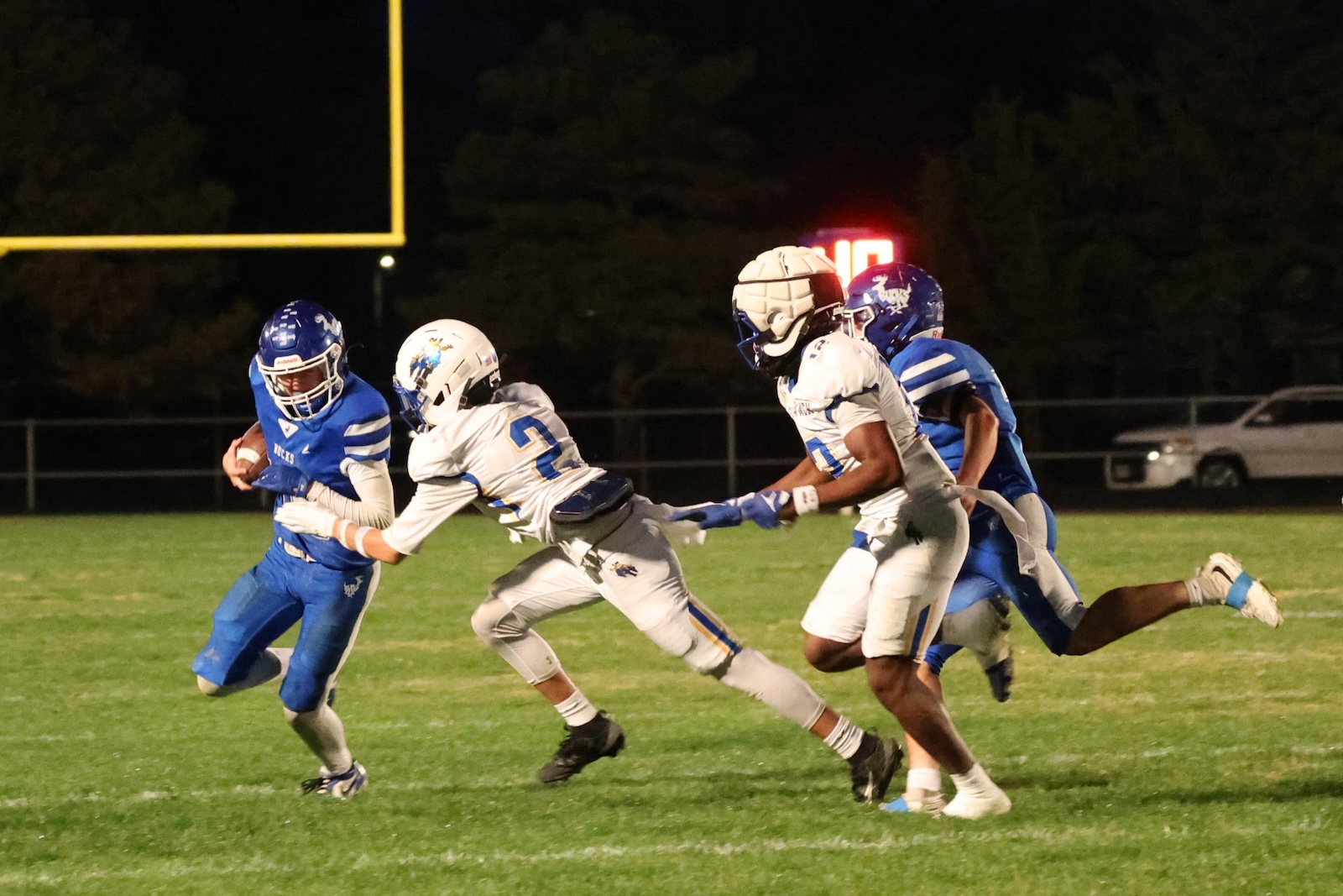 Football player in a blue jersey runs with the ball while white-clad defenders chase during a night game.