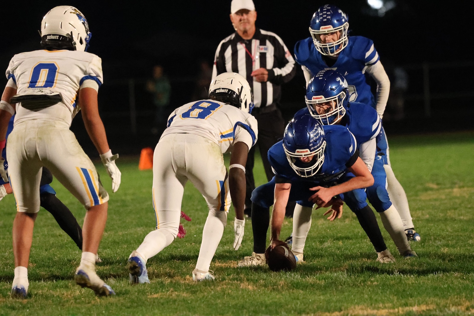 Youth football players in blue and white uniforms crouch around a loose ball on a grassy night field.