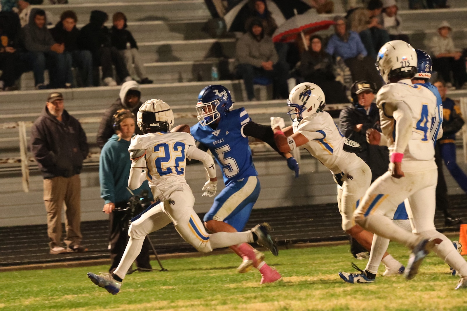 Blue team ball carrier (number 5) runs past a white defender (number 22) toward the end zone as teammates pursue. Spectators watch from stands in the background.
