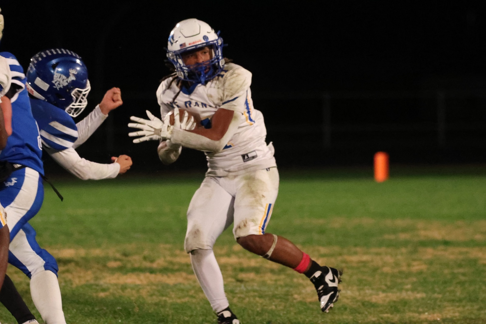 American football player in a white uniform runs with the ball while a defender in blue closes in during a night game on a grassy field, an orange pylon visible in the background.