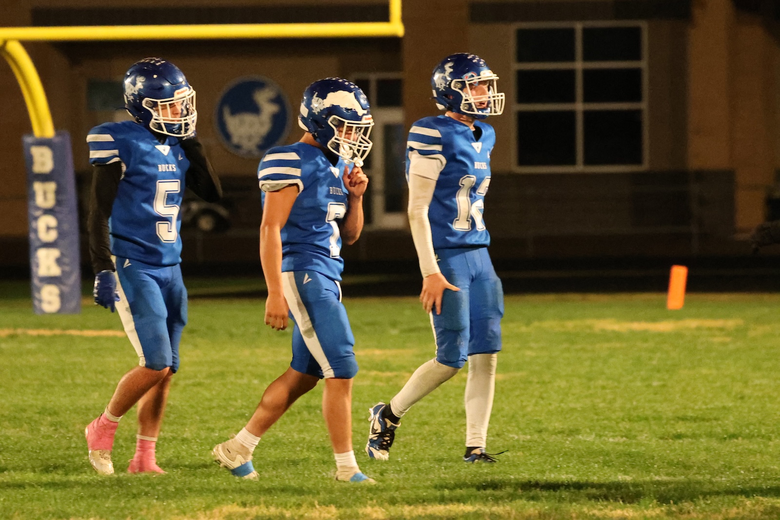Three football players in blue jerseys and white helmets walk across a darkened field at night, near a yellow goalpost padding and an orange pylon in the background.