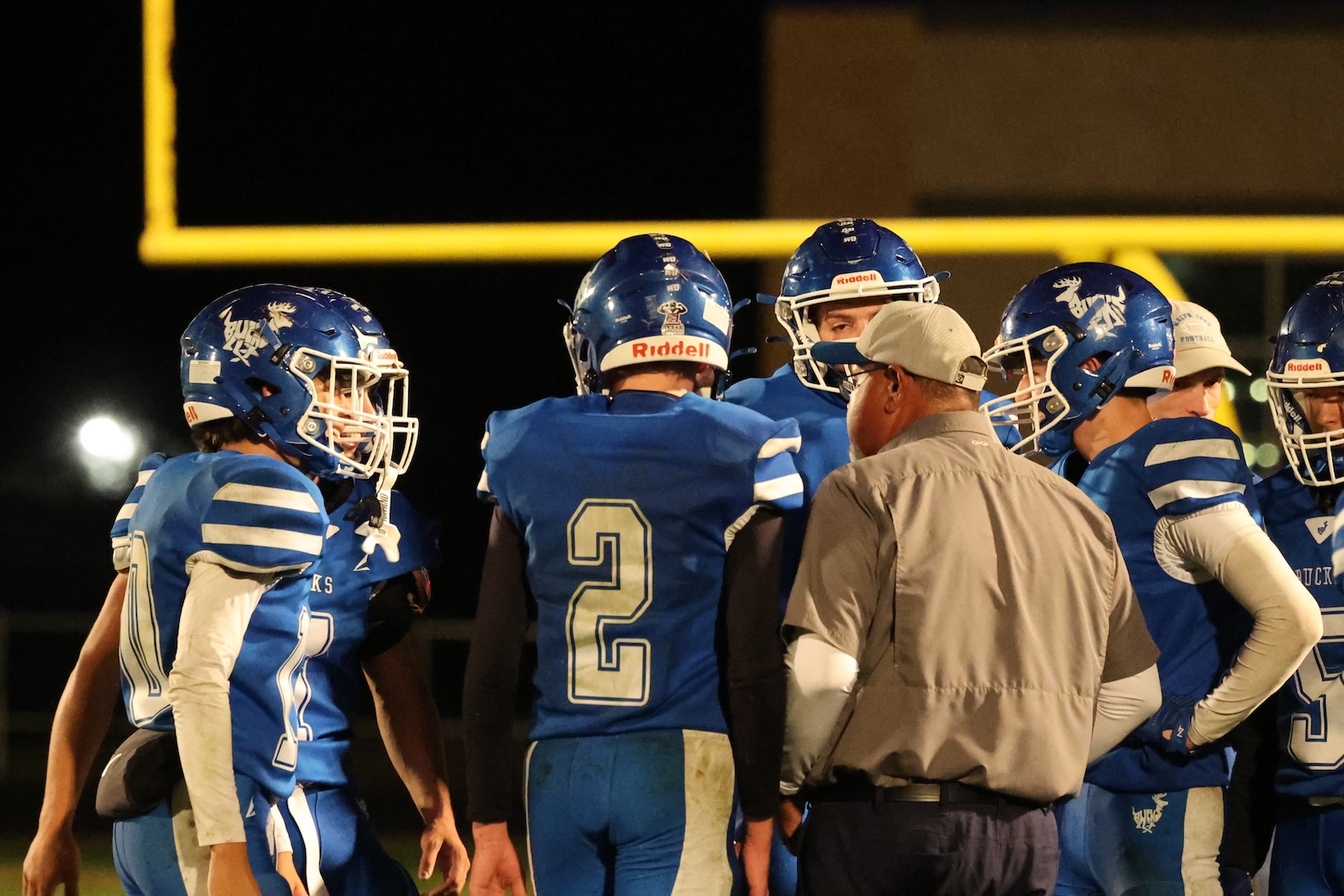 Nighttime football huddle: players in blue uniforms and helmets circle around a coach on the field near a yellow goal post.