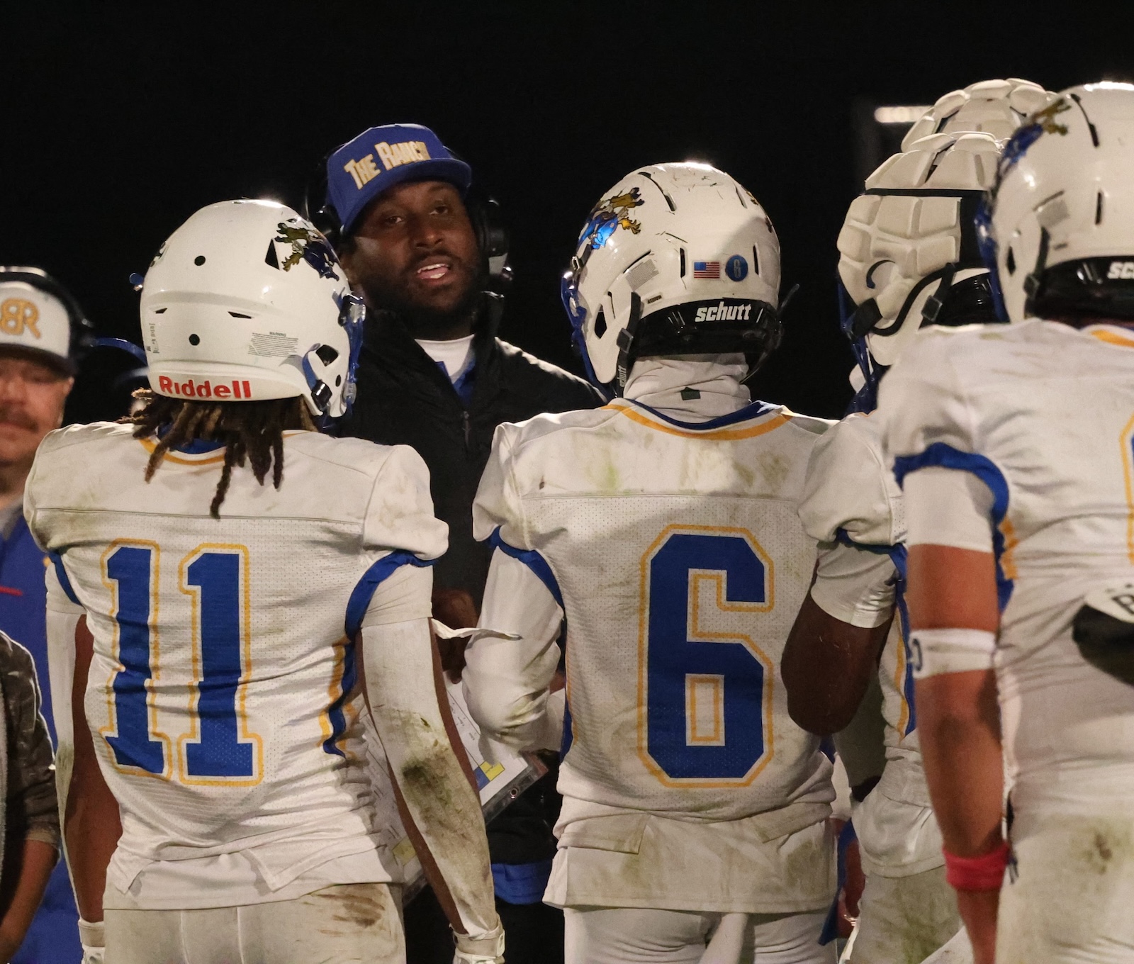 Football players in white jerseys huddle around their coach at night; numbers 11 and 6 visible on their backs.