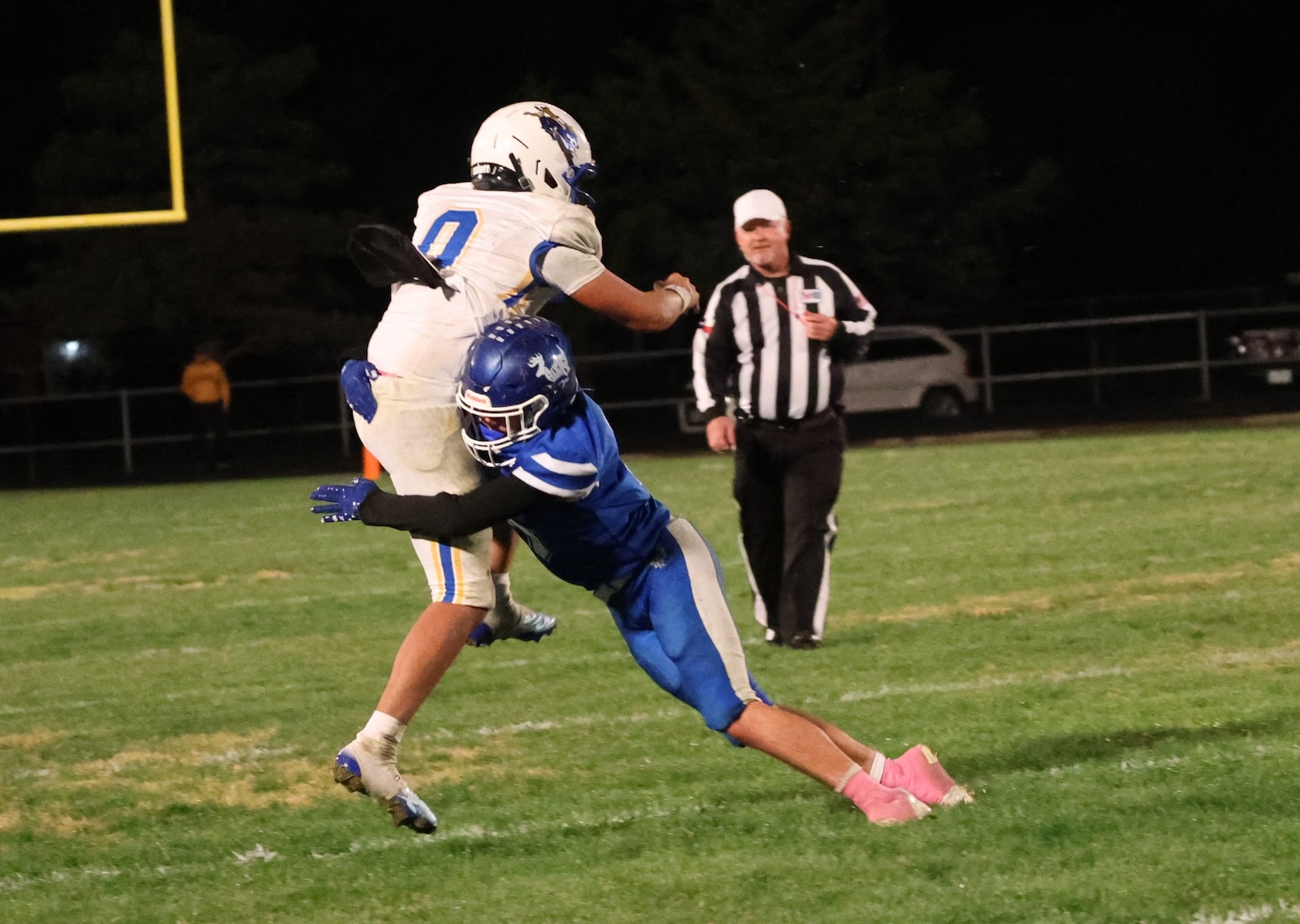 Nighttime football play: a blue-numbered defender tackles a white-uniform runner as they go airborne on a grassy field. A striped referee watches in the background.