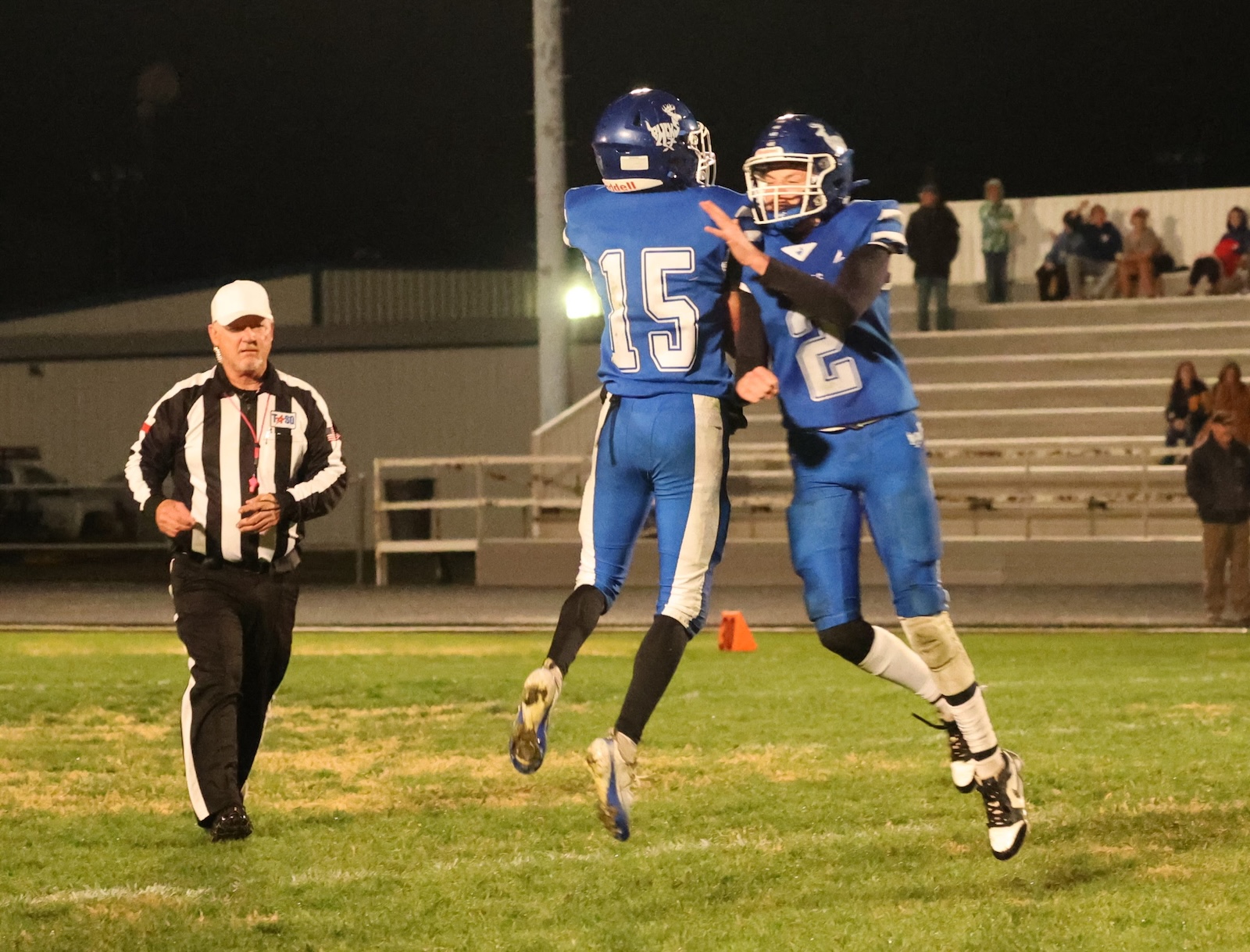 Two blue-jersey football players jump and celebrate on a grassy field as a referee approaches left. Fans in bleachers in the background at night.
