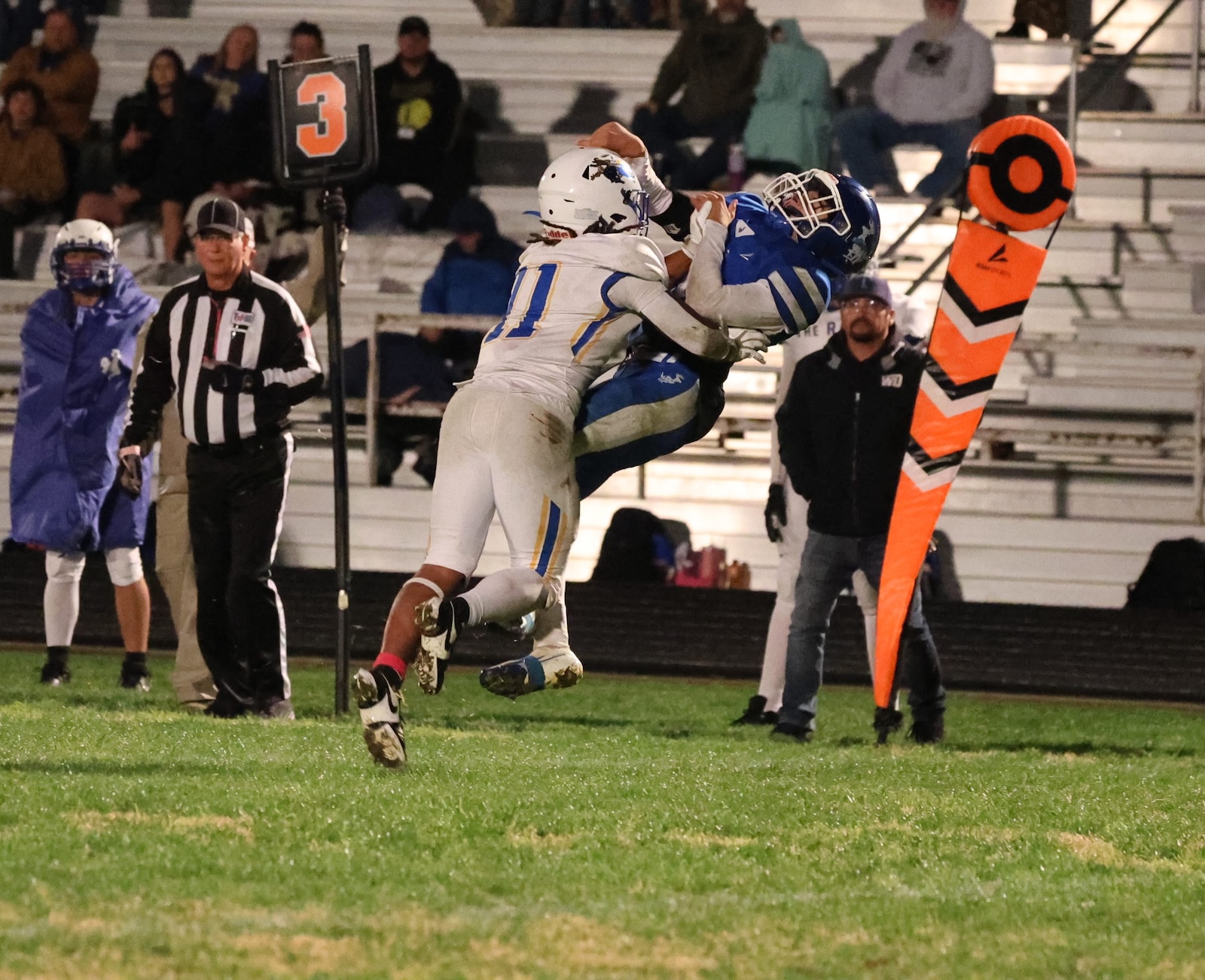 Two football players in white and blue jerseys collide mid-air near the sideline while an official watches from the left; an orange pylon marks the end zone nearby.